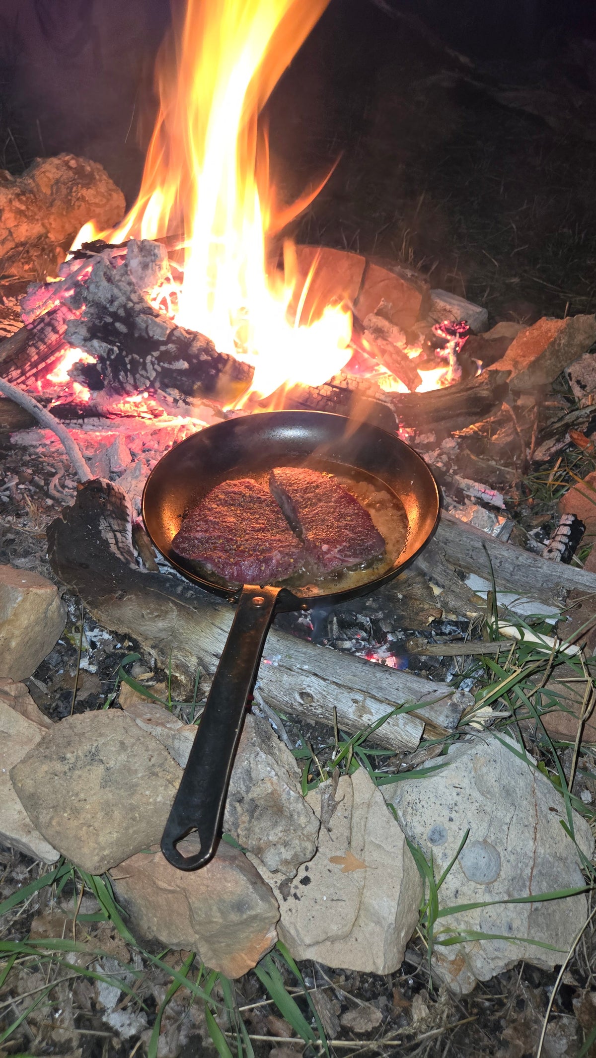 Steak searing in The Original carbon steel skillet set on the coals of a campfire at night