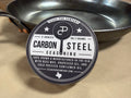 Carbon Steel Seasoning tin, closed, leaning against a carbon steel skillet