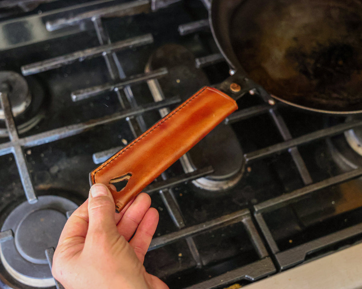 A hand holding The Original carbon steel skillet by its leather-covered handle over a gas range