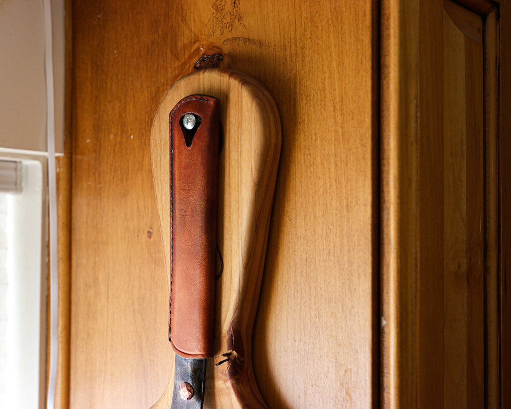The Original carbon steel skillet hanging on a wooden board with the Leather Handle Cover on its handle
