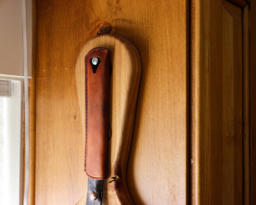 The Original carbon steel skillet hanging on a wooden board with the Leather Handle Cover on its handle