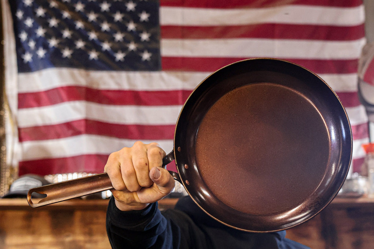 A hand holding a Texas Pan Company carbon steel skillet in front of an American flag