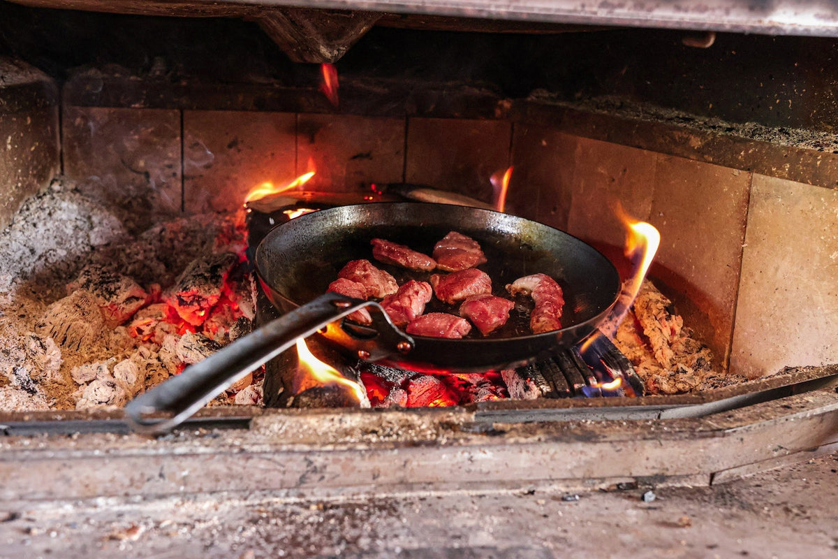 The Original carbon steel skillet searing steak on the coals of a wood fire