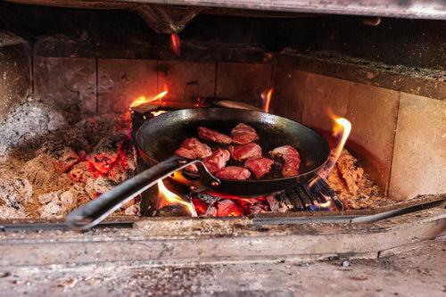The Original carbon steel skillet searing steak on the coals of a wood fire