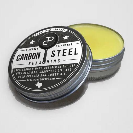 Carbon Steel Seasoning tin with the lid leaning against it, showing the pale yellow balm inside