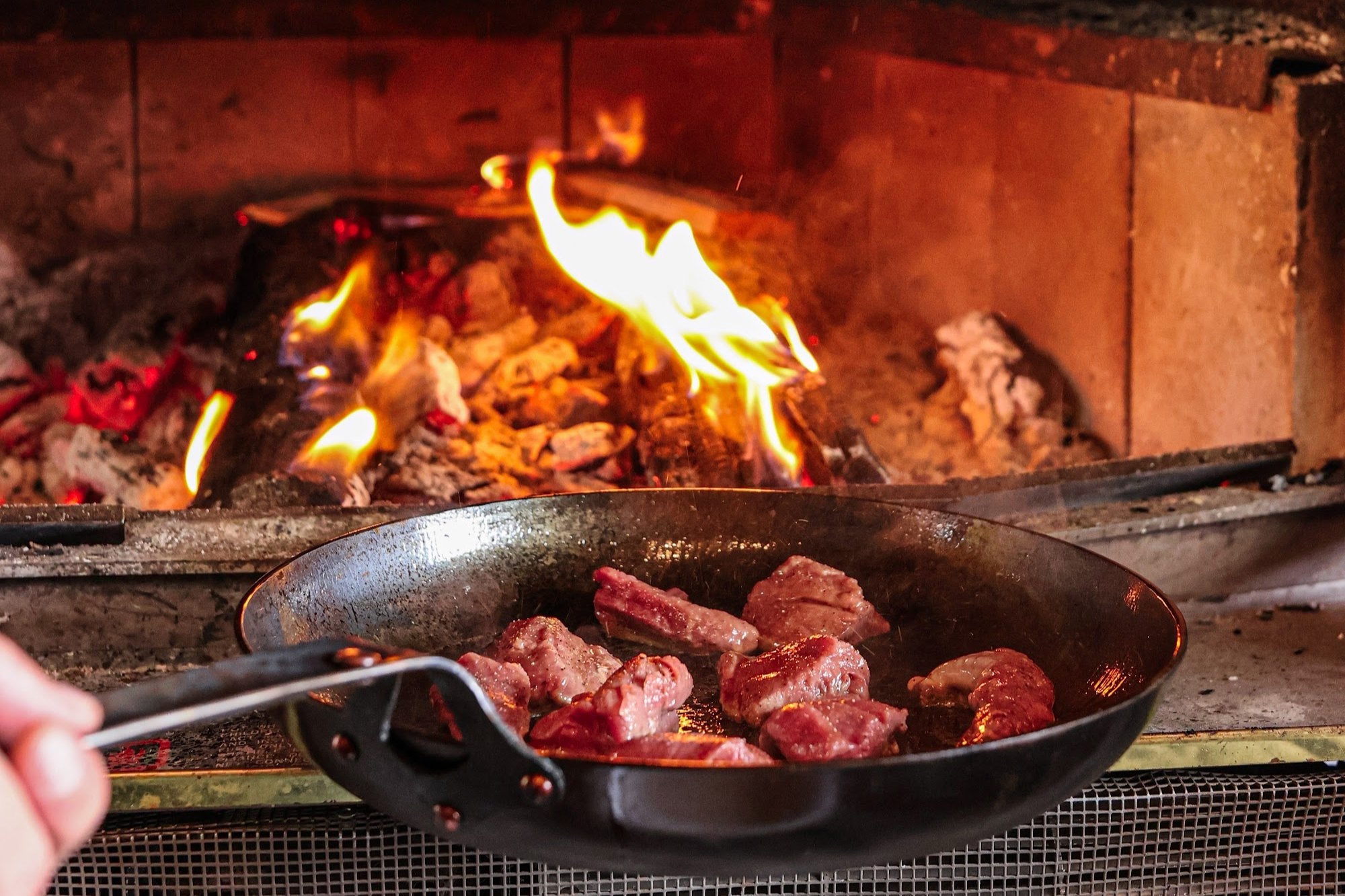 A hand holding The Original carbon steel skillet by the handle over a wood fire
